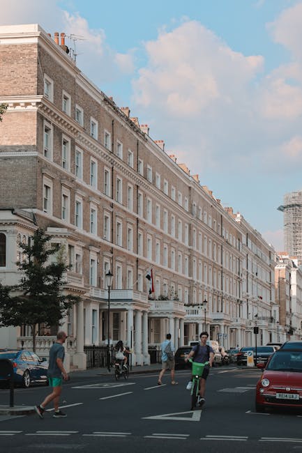 The image shows a row of multi-storey Victorian-style residential buildings with beige brick facades and white window frames along a busy street in South Kensington. In the foreground, pedestrians walk across the road, including a cyclist riding a bicycle wearing a green top and black shorts. To the right, a red car is parked on the street, while other vehicles are parked along the curb. The building's ground floors feature classic architectural details such as columns and balcony railings. Some of the upper windows have small chimneys extending from the roofline. The scene is well-lit with natural daylight, and a clear blue sky with light clouds is visible above. The image captures a typical urban environment where house removals and furniture transport might occur as part of a home relocation, with the proximity of the pavement, street, and residential buildings indicating a central London location. Occasionally, Movers South Kensington may be involved in providing removal services in such environments, including loading and logistical planning during house moves. In the background, a construction scaffolding structure indicates ongoing building work nearby.