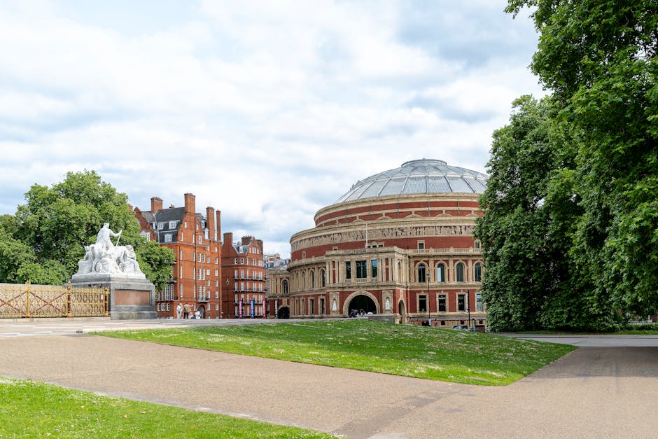 A wide view of a city park showing a large, circular, historic building with a domed roof and detailed brickwork, situated behind a well-maintained grassy area and paved pathways. To the left, a white marble statue atop a pedestal, depicting classical figures, is partially obscured by green trees that line the park's perimeter. The sky above is partly cloudy with patches of blue, and in the background, red-brick residential buildings with multiple windows and chimneys are visible. The scene captures a moment of activity with a few people walking near the building, reflecting a typical urban environment where house removals or furniture transport might be coordinated. The natural lighting highlights the textures of the building's architecture and the lush greenery, aligning with residential and commercial moving and packing processes managed by companies like Movers South Kensington.