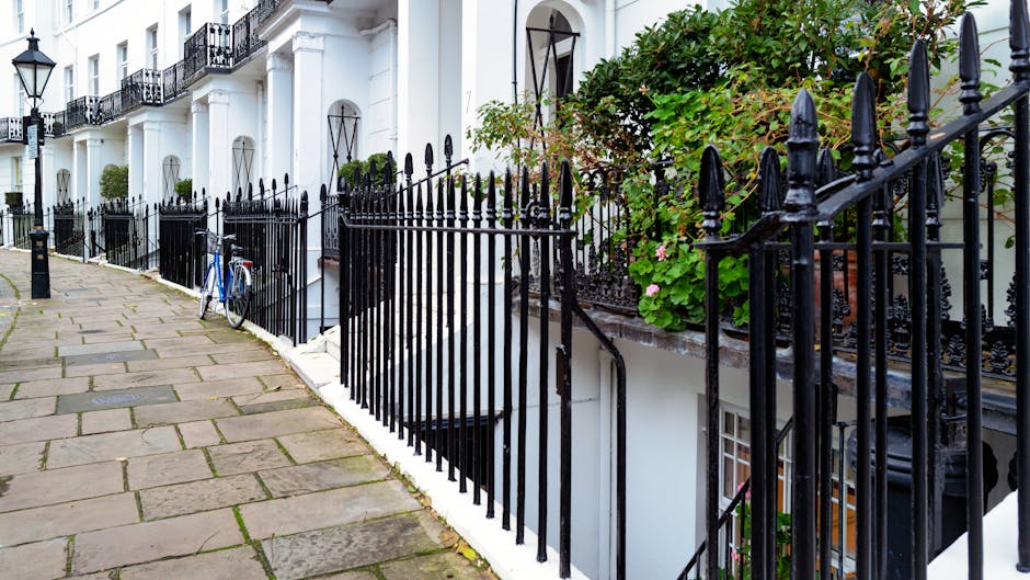 Image depicting the exterior of a white Victorian-style terraced house with black wrought iron railings along a paved sidewalk, located in South Kensington. A blue bicycle is leaning against the fence near the front of the property. The entrance features a small staircase with black metal railings, leading down to a basement door, which is partially visible beneath the upper-level windows. Green foliage and flowering plants grow around the front steps, adding colour to the scene. The fence and house structure suggest a typical London residential street, with natural daylight illuminating the scene, indicating daytime. This setting is relevant to house removals and home relocation services provided by Movers South Kensington, especially in the context of moving logistics or packing and loading processes associated with residential moves.