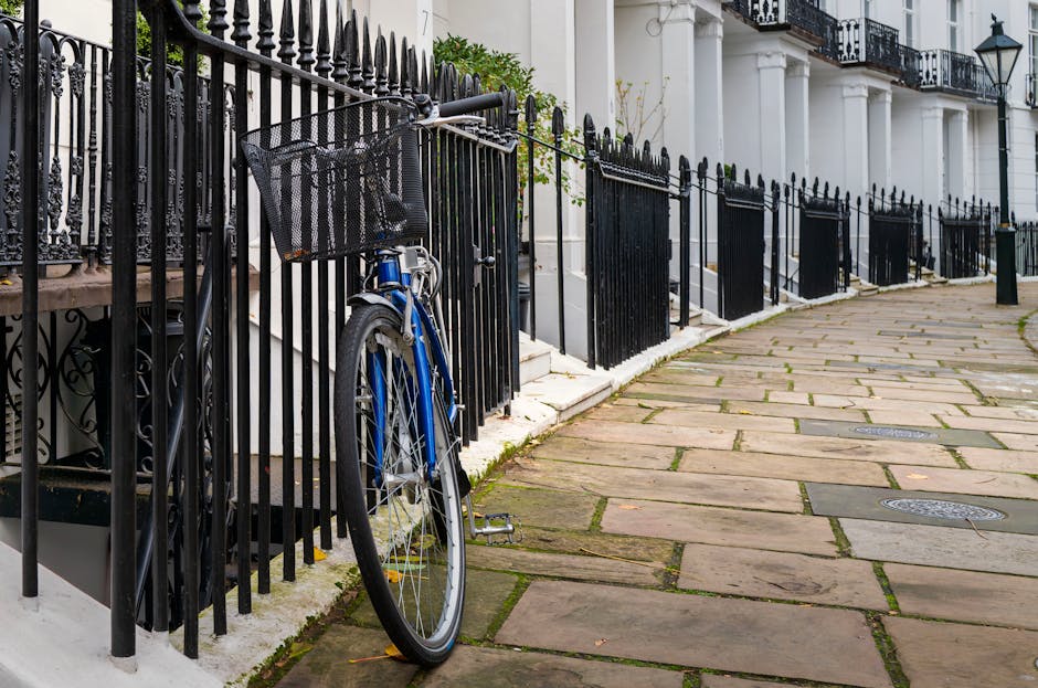 A wide view of a classic four-storey residential building on a street in South Kensington, London, with ornate cream-coloured facades, decorative cornices, and sash windows. The building's ground floor features white columns supporting a balcony above the entrance, and a black fence surrounds the front steps. Several pedestrians walk along the pavement, and a cyclist is seen riding past the building. parked cars line the street, including a red vehicle in the foreground. The sky above is mostly clear with some scattered clouds, and a construction scaffolding is visible on a nearby building. This scene reflects typical urban residential architecture suitable for house removals and furniture transport, as provided by Movers South Kensington, and illustrates a busy day involving moving or packing activities in an area requiring permits for residential relocations.
