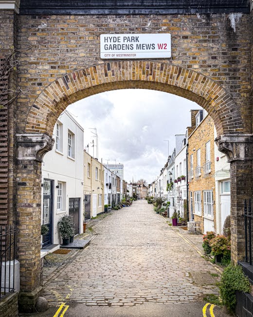 A view through a brick archway showing a narrow cobblestone residential street in Gloucester Road mews, South Kensington, with closely packed terraced houses on both sides. The houses have white and cream facades with small front gardens, some with potted plants and hedges. The street extends into the distance under a cloudy sky. Visible at the top of the arch is a white street sign reading 'Hyde Park Gardens Mews W2' with a note indicating it is part of the City of Westminster. In the context of a house removal, this image depicts the typical tight access conditions that Movers South Kensington might encounter when planning furniture transport and packing for a home relocation in such historic mews streets, highlighting the challenges of narrow entrances and limited space for vehicle access during a professional removals process.