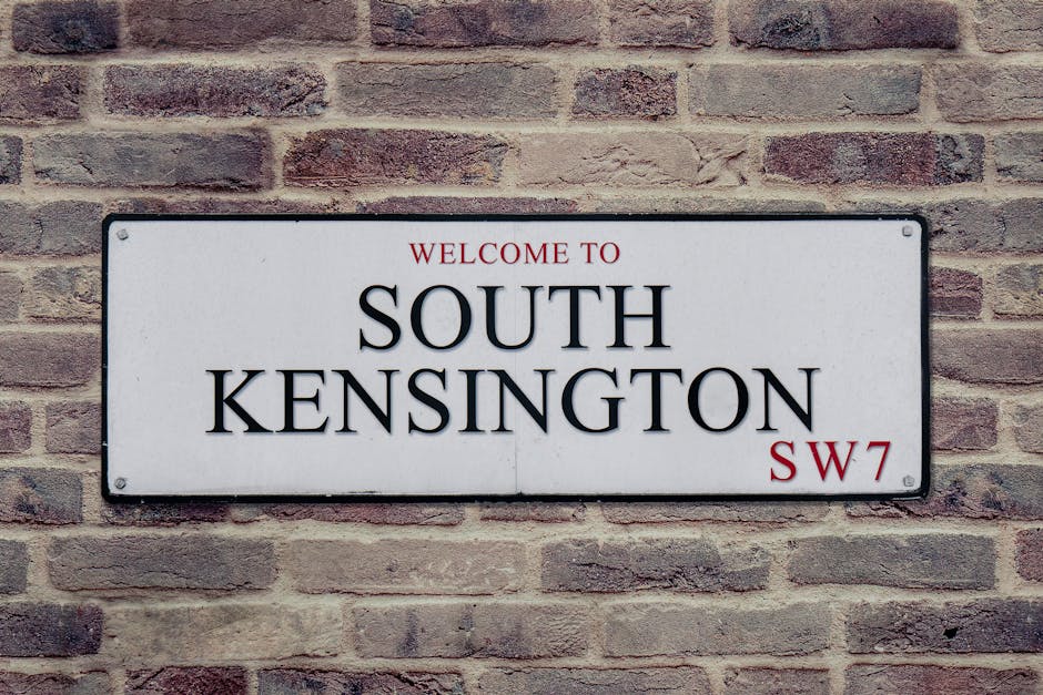 A rectangular white street sign affixed to a red brick wall displays the words 'Welcome to South Kensington SW7' in bold black and red lettering, with 'South Kensington' prominently centered. The sign is mounted with four screws, one at each corner. The brick wall behind the sign consists of evenly laid bricks with a slightly weathered appearance, showcasing various shades of reddish-brown and beige. This setting is typical of the exterior of a residential or commercial property in South Kensington, London. The sign’s clarity and placement suggest it marks the entrance to a neighborhood, contributing to local wayfinding. The scene is evenly lit, with no shadows, indicating natural daylight. Such signage is common in areas requiring clear identification for visitors, and it reflects the context of home relocation or furniture transport services operating in this part of Gloucester Road mews, where space constraints may pose challenges during packing and loading processes handled by [COMPANY_NAME].