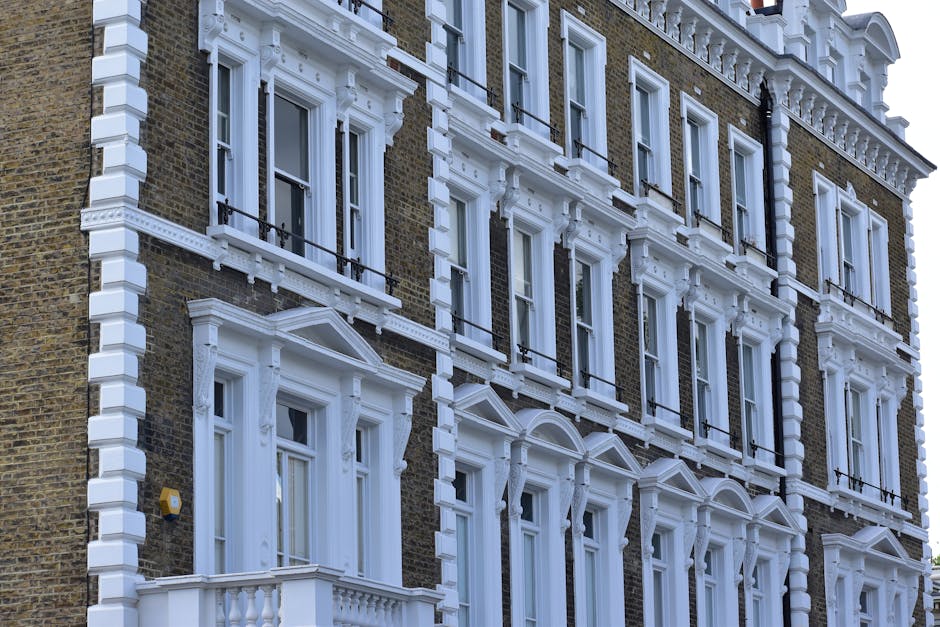 A multi-storey historic residential building with a brick facade and ornate white decorative window frames and mouldings. The windows are double-glazed with small black metal railings in front. The lower windows have small balcony balustrades, while the upper windows are sash style. The building's exterior features white painted architectural details, including cornices and column-like embellishments around the windows. External lighting fixtures are visible on the facade. The scene appears to be in daylight, and the building is likely located in a residential area suitable for house relocations or removals services by Movers South Kensington, who specialise in home moving logistics around Earls Court, South Kensington, and similar areas.