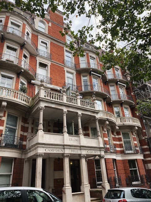 A multi-storey residential building with a classic architectural style, featuring a white stone façade with decorative columns and balustrades on the entrance terrace, situated at 139-36 Campden Hill Court. The building has red brickwork and white window frames, with several balconies enclosed by ornate black wrought iron railings. The image was taken from the street, with a view of the building partly obscured by green leafy trees overhead. In the foreground, parked cars are visible on the pavement, indicating an urban area typical for house removals and furniture transport in Earls Court and South Kensington. The scene reflects a typical setting for a professional house removal involving careful handling of furniture and packed boxes within this historic residential property, as part of home relocation services by Movers South Kensington.