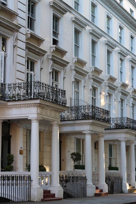 A multi-storey historic residential building with a brick facade and ornate white decorative window frames and mouldings. The windows are double-glazed with small black metal railings in front. The lower windows have small balcony balustrades, while the upper windows are sash style. The building's exterior features white painted architectural details, including cornices and column-like embellishments around the windows. External lighting fixtures are visible on the facade. The scene appears to be in daylight, and the building is likely located in a residential area suitable for house relocations or removals services by Movers South Kensington, who specialise in home moving logistics around Earls Court, South Kensington, and similar areas.