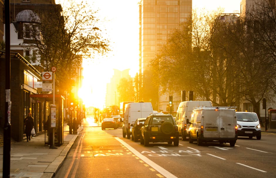 A busy urban street scene during sunset in South Kensington with traffic congestion involving several vehicles including white vans, small cars, and a delivery truck, lining both sides of the road. The pavement on the left side is occupied by pedestrians walking past a row of shops and a bus stop. A public transport sign is visible near the bus shelter, which features a glass enclosure. Tall buildings and leafless trees line the street, with modern high-rise structures visible in the background, bathed in warm, golden sunlight. The light reflects off the vehicles and the shiny surfaces of the shop windows, creating a vibrant yet congested atmosphere typical of city logistics and home relocation activities handled by Movers South Kensington, especially during peak moving times or delays in SW7, South Kensington.