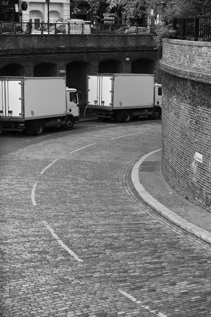 A black and white photograph showing a curved, cobbled driveway partially enclosed by a brick wall with a small sign attached to it. Two large white moving trucks are parked along the driveway, with their rear doors facing away from the camera. Inside the trucks, visible partially through open doors, are packing materials such as cardboard boxes, plastic crates, and wrapped furniture. Some of these items are on dollies and served with blankets and straps for secure transportation. The driveway is bordered by a metal railing and a sidewalk with a brick wall that curves around the corner, leading to the entrance of a residential or commercial building. In the background, there are trees and buildings with awnings, suggesting an urban setting in South Kensington, where house removals and furniture transport are commonly coordinated by Movers South Kensington as part of a home relocation process. The scene captures the loading process typical of professional removals services focused on packing and moving tasks, with natural lighting highlighting the materials and equipment used in the move.