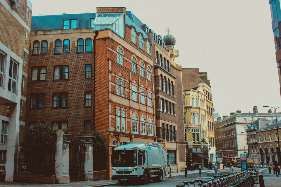 A busy urban street scene during sunset in South Kensington with traffic congestion involving several vehicles including white vans, small cars, and a delivery truck, lining both sides of the road. The pavement on the left side is occupied by pedestrians walking past a row of shops and a bus stop. A public transport sign is visible near the bus shelter, which features a glass enclosure. Tall buildings and leafless trees line the street, with modern high-rise structures visible in the background, bathed in warm, golden sunlight. The light reflects off the vehicles and the shiny surfaces of the shop windows, creating a vibrant yet congested atmosphere typical of city logistics and home relocation activities handled by Movers South Kensington, especially during peak moving times or delays in SW7, South Kensington.