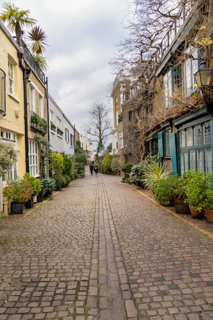 A cobbled residential street with natural stone paving, flanked by multi-storey buildings on both sides. On the left, there are four-story houses with cream and white facades, featuring large windows and small balconies, some with potted plants. On the right, a mix of older and more modern buildings with metal staircases, glass panels, and outdoor plants in pots and planters. Sparse leafless trees are visible alongside the buildings, with some greenery and shrubs near the base of the houses. The sky is overcast, and the street appears to be quiet with a few pedestrians walking in the distance. This setting suggests a typical urban neighbourhood that could be involved in house removals or relocations, with movers potentially using the street for loading and unloading furniture and boxes during a home relocation, such as furniture transport or packing and moving activities, possibly coordinated by Movers South Kensington.
