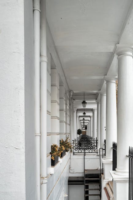 A long, narrow exterior corridor of a residential building with white-painted walls, featuring large round white columns supporting the ceiling. Several black wrought-iron fences and gates are positioned along the walkway, separating small steps or entryways. Potted plants with dried or brown foliage are placed on the ledges along the corridor. The ceiling has hanging black lantern-style light fixtures, and the area appears well-lit by natural daylight. The scene is typical of a townhouse or Victorian-style terrace, with clean lines and traditional architectural details, suitable for home relocation or furniture transport services such as those offered by Movers South Kensington in the context of moving preparations near South Kensington station.