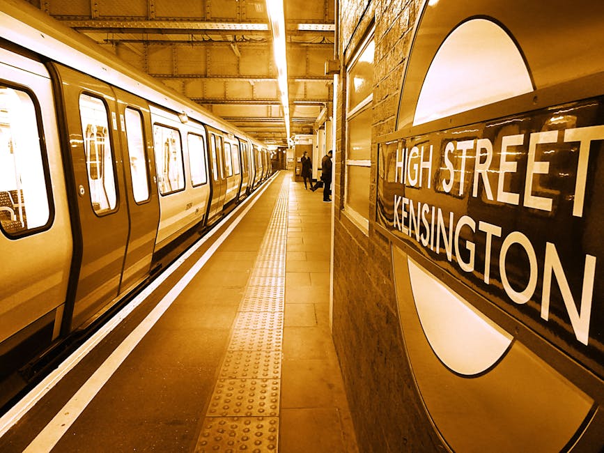 Inside a London Underground station at South Kensington, showing a platform with a yellow subway train parked alongside. The train has multiple doors with windows, and the platform is tiled with tactile paving strips near the edge for safety. The station's brick wall features a large roundels sign with the words 'HIGH STREET KENSINGTON,' and a few passengers are visible in the background, some walking or standing. Overhead lighting illuminates the scene, casting a warm glow. As part of a home relocation or furniture transport process, the image depicts the loading area where goods such as boxes, furniture wrapped in plastic or blankets, and moving equipment like trolleys or straps could be present for efficient packing and loading onto the train, consistent with moving services offered by Movers South Kensington.