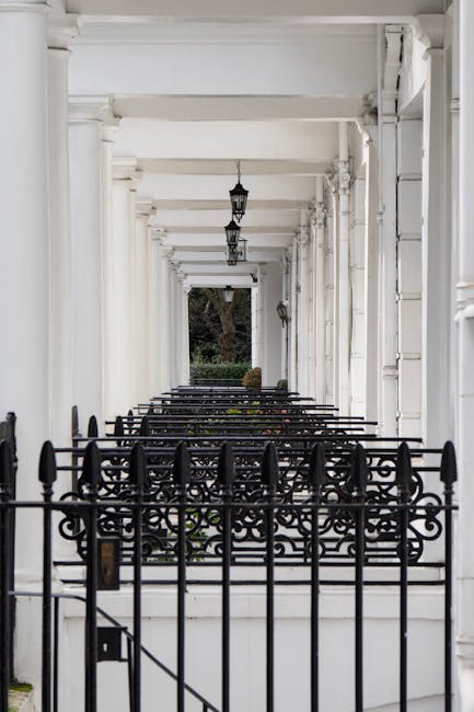 A long, narrow exterior corridor of a residential building with white-painted walls, featuring large round white columns supporting the ceiling. Several black wrought-iron fences and gates are positioned along the walkway, separating small steps or entryways. Potted plants with dried or brown foliage are placed on the ledges along the corridor. The ceiling has hanging black lantern-style light fixtures, and the area appears well-lit by natural daylight. The scene is typical of a townhouse or Victorian-style terrace, with clean lines and traditional architectural details, suitable for home relocation or furniture transport services such as those offered by Movers South Kensington in the context of moving preparations near South Kensington station.